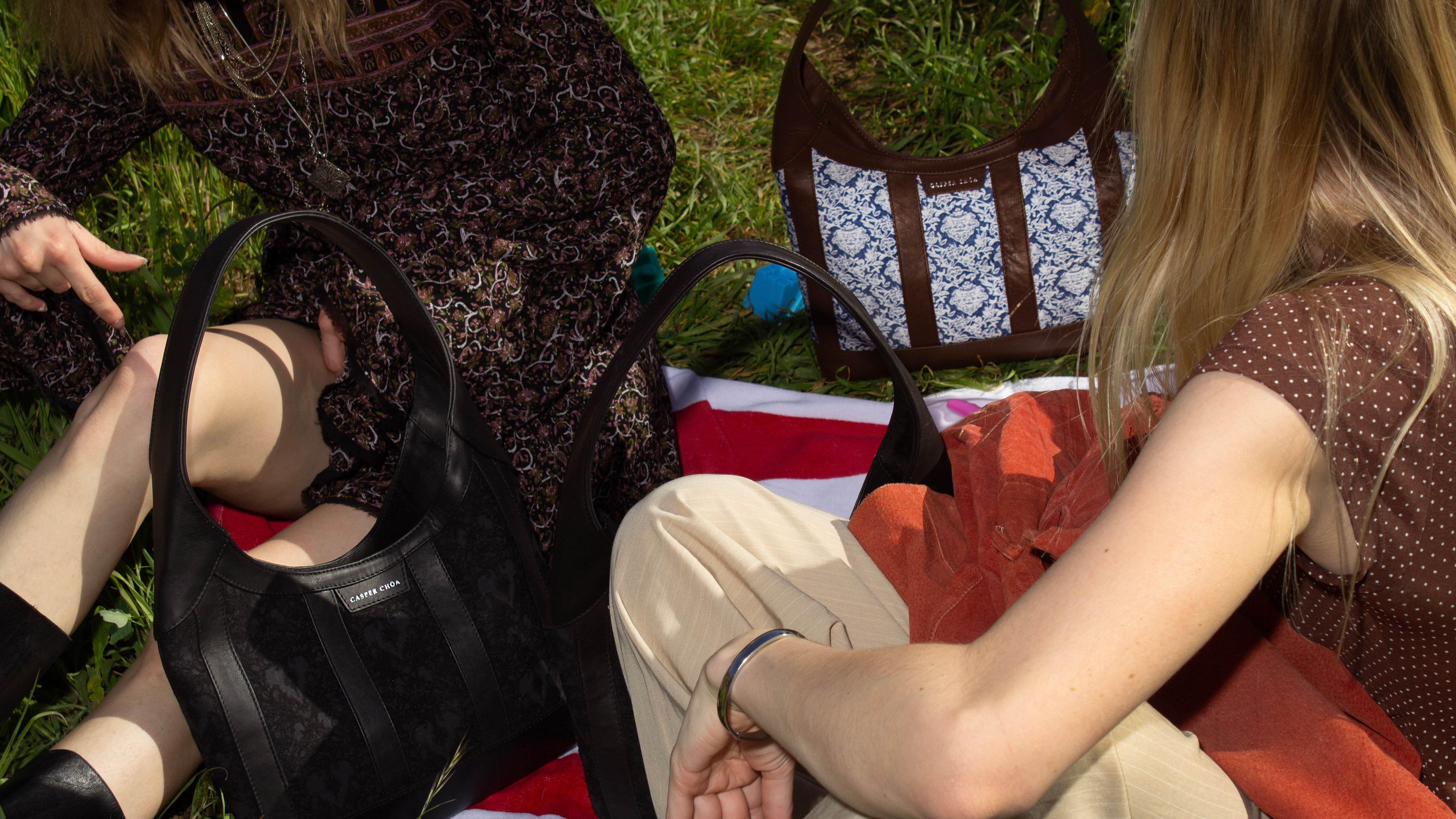 two women sitting next to handbags on the grass