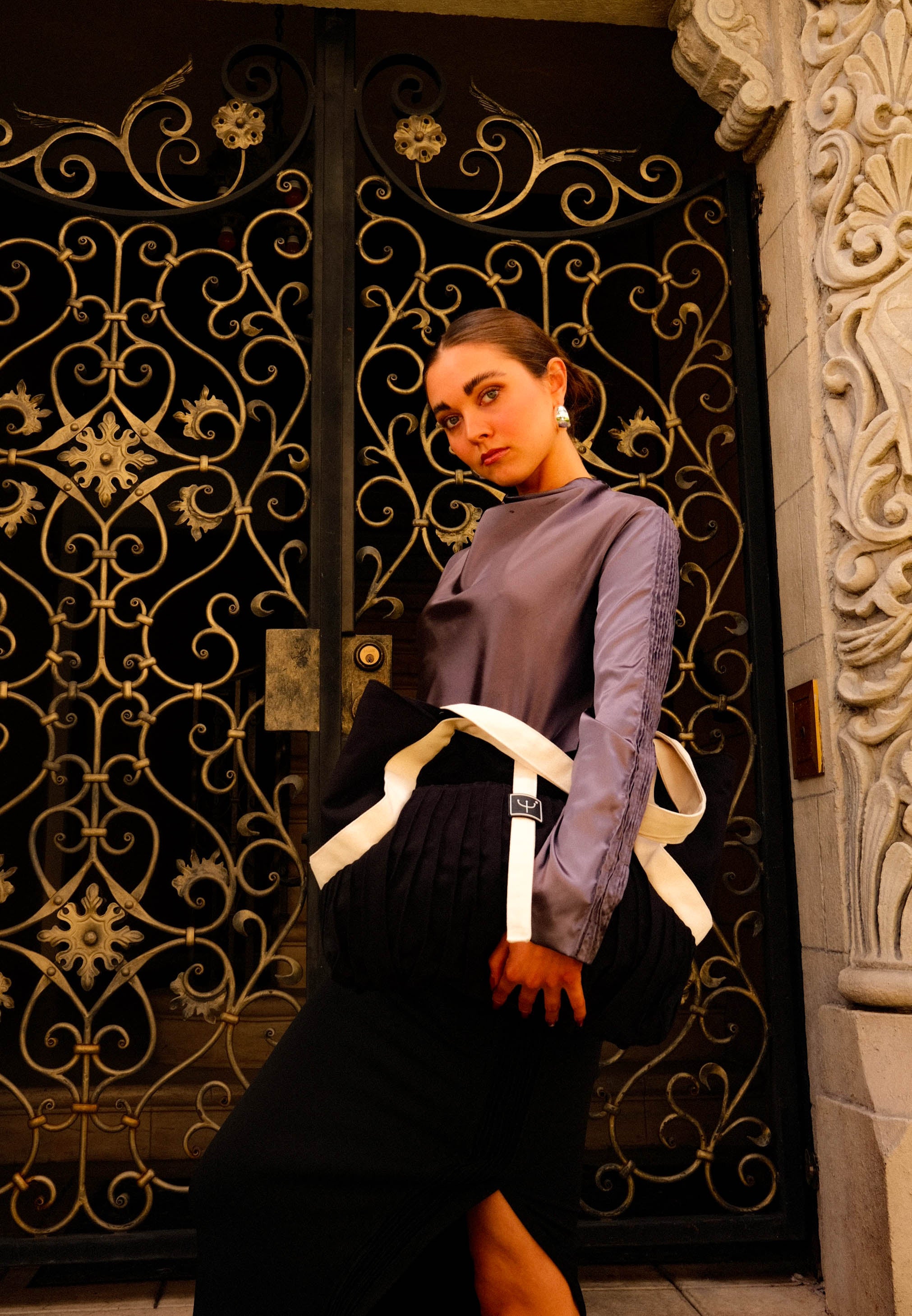 picture of woman in purple long sleeved top and black maxi skirt holding a black and white tote bag in front of iron door