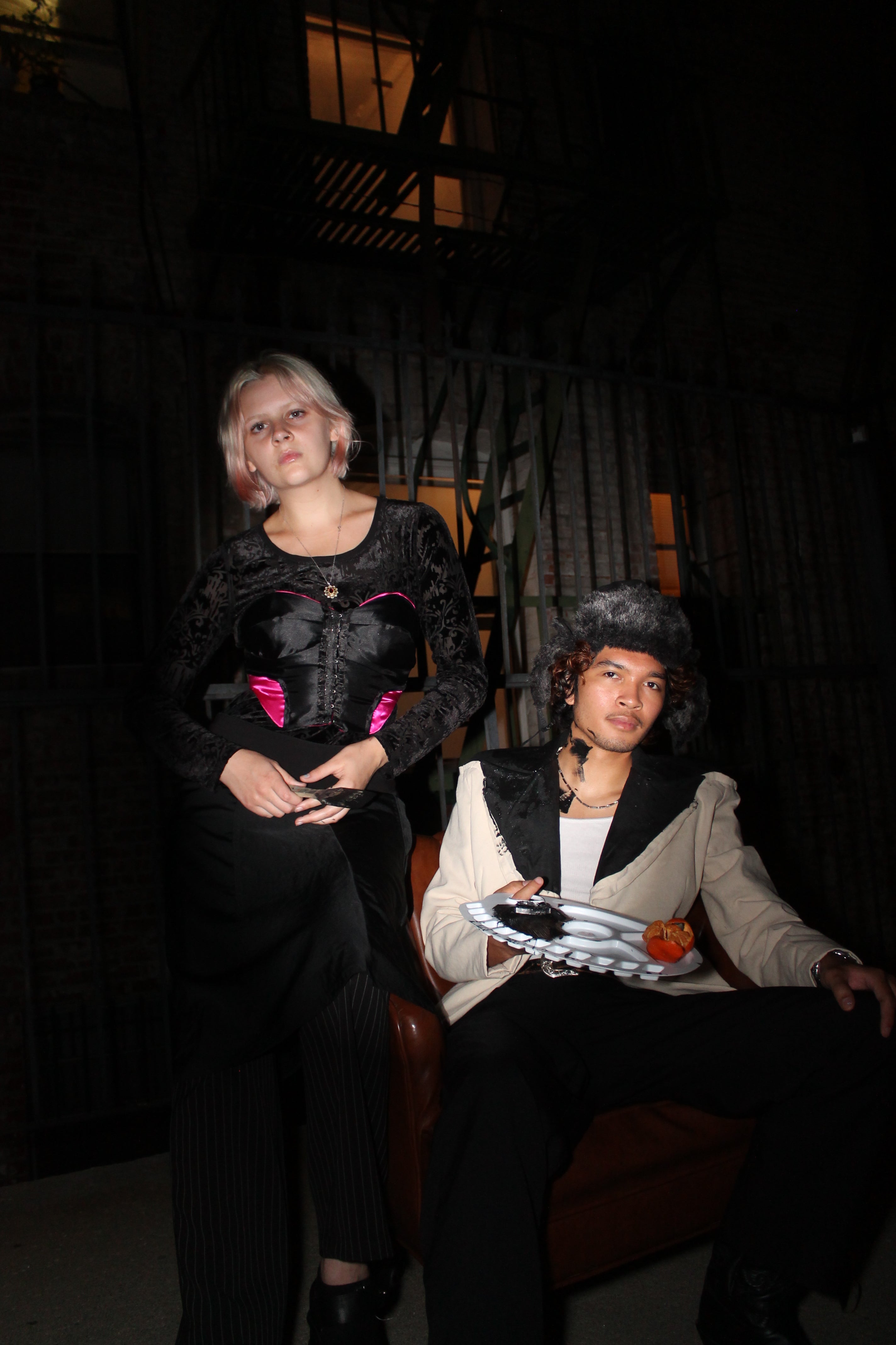 picture of man in winter hat, ivory and black avant garde blazer, white tank top, and black pants with a woman in a black and pink bustier, black long sleeved shirt, black skirt, and black pants in an alley