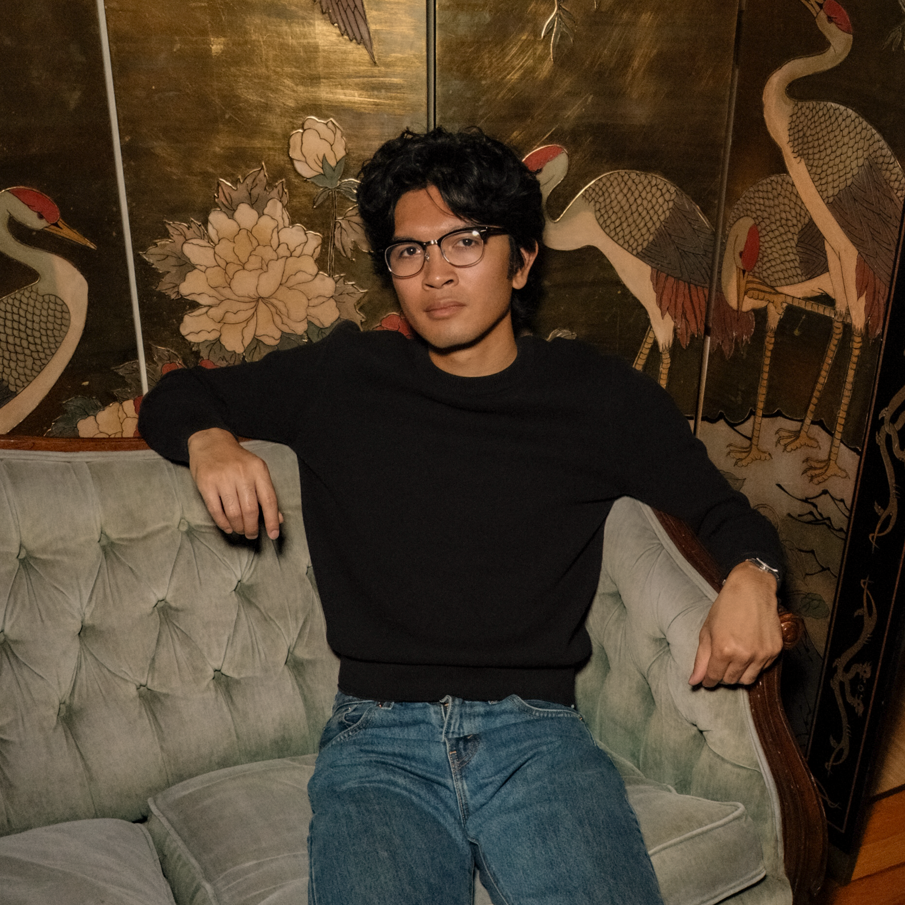 Casper Choa sitting on a vintage couch with decorative gold room divider in the background