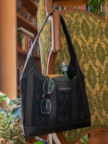 Eyeglasses, books, and glass bottle in a black leather handbag with black/black heart-patterned jacquard on a chair with a bookshelf background
