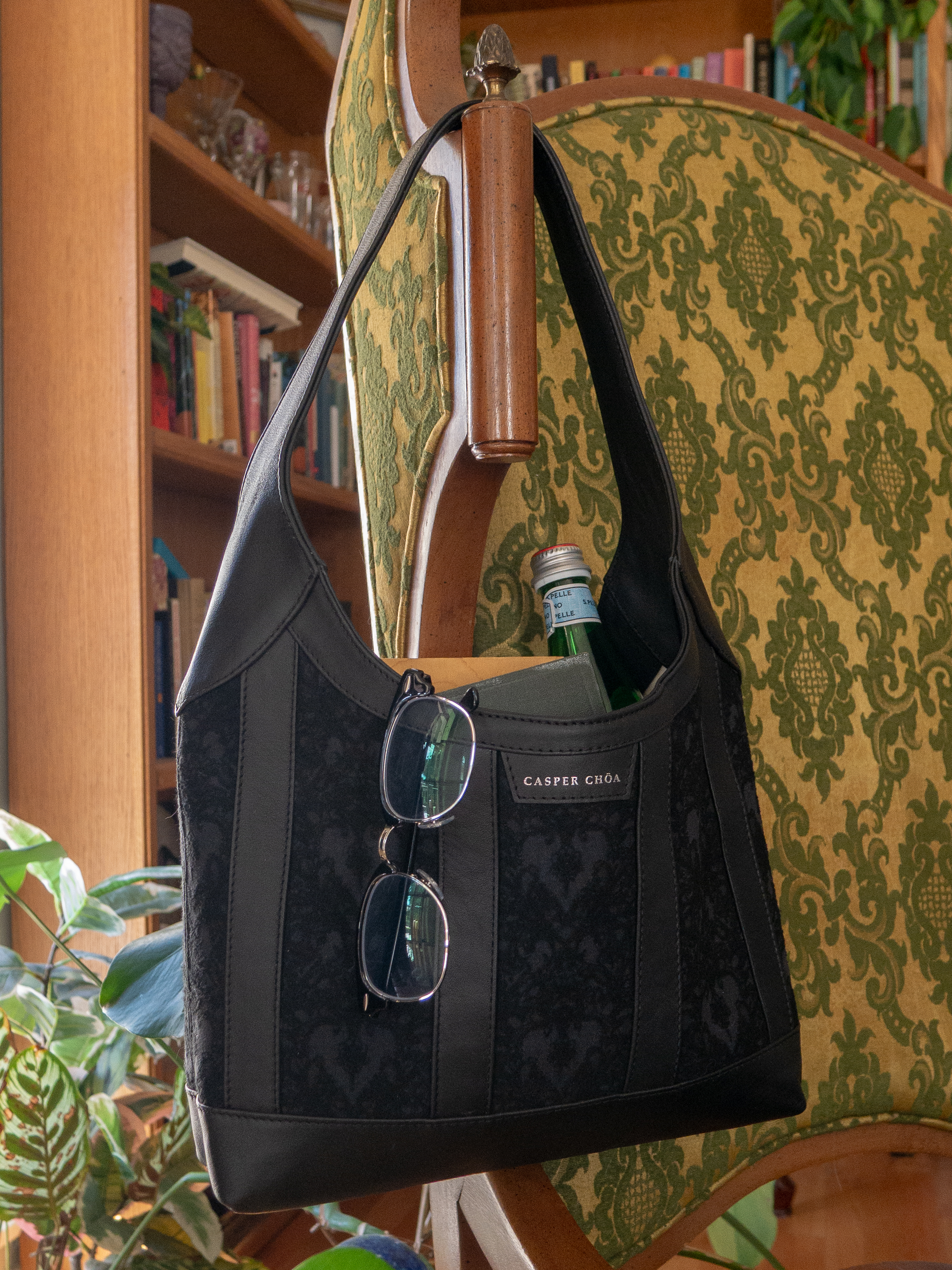 Eyeglasses, books, and glass bottle in a black leather handbag with black/black heart-patterned jacquard on a chair with a bookshelf background