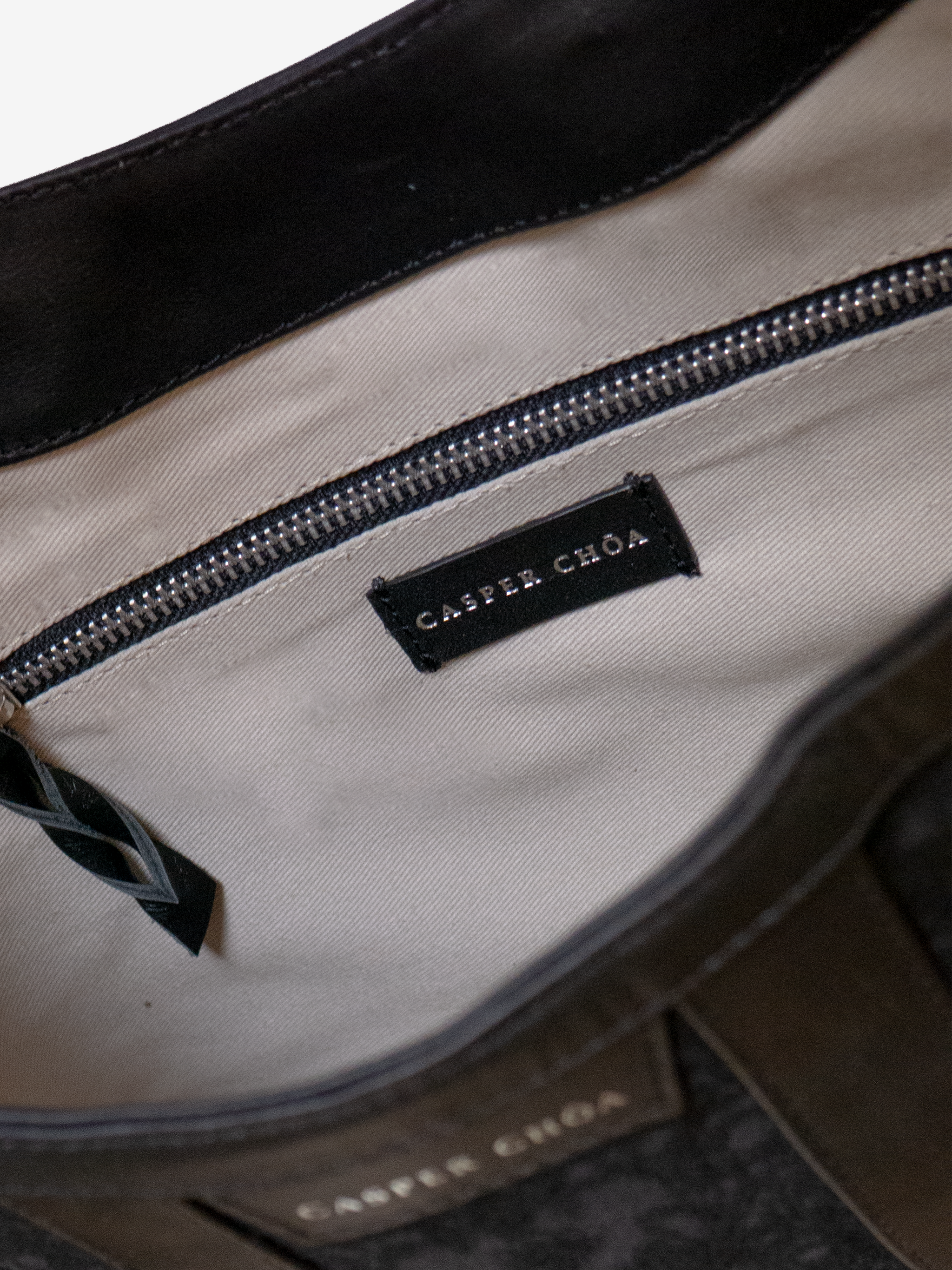 Close-up of a beige twill interior lining with a silver-tone CASPER CHOA stamped signature on black calfskin and a silver zipper with black calfskin zipper pull