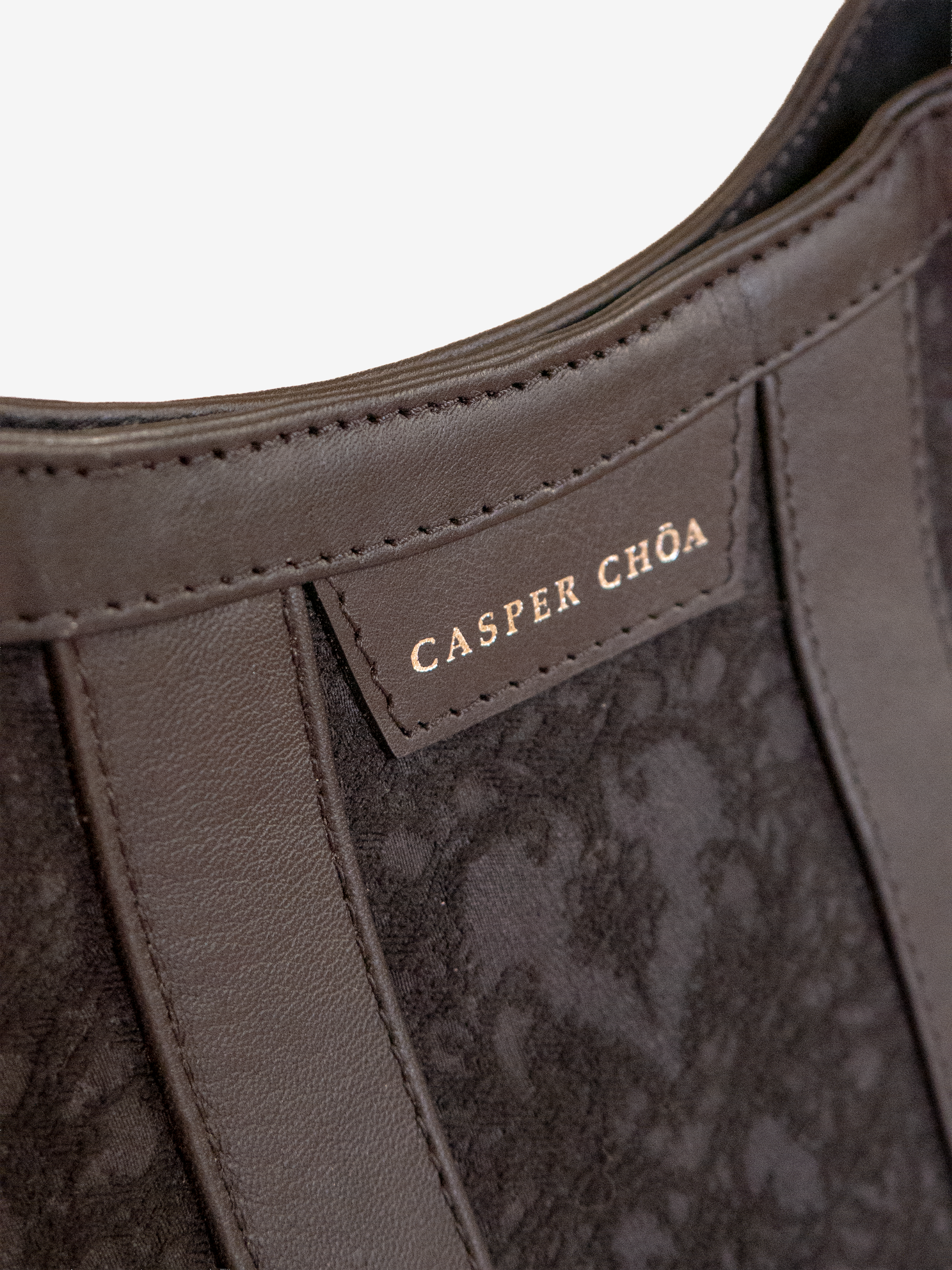 Close-up of a silver-tone CASPER CHOA stamped signature on a back leather handbag with black/black heart-patterned jacquard on a white background