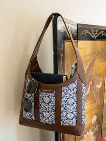 Sunglasses, books, and glass bottle in a brown leather handbag with blue/white floral jacquard against a decorative room divider