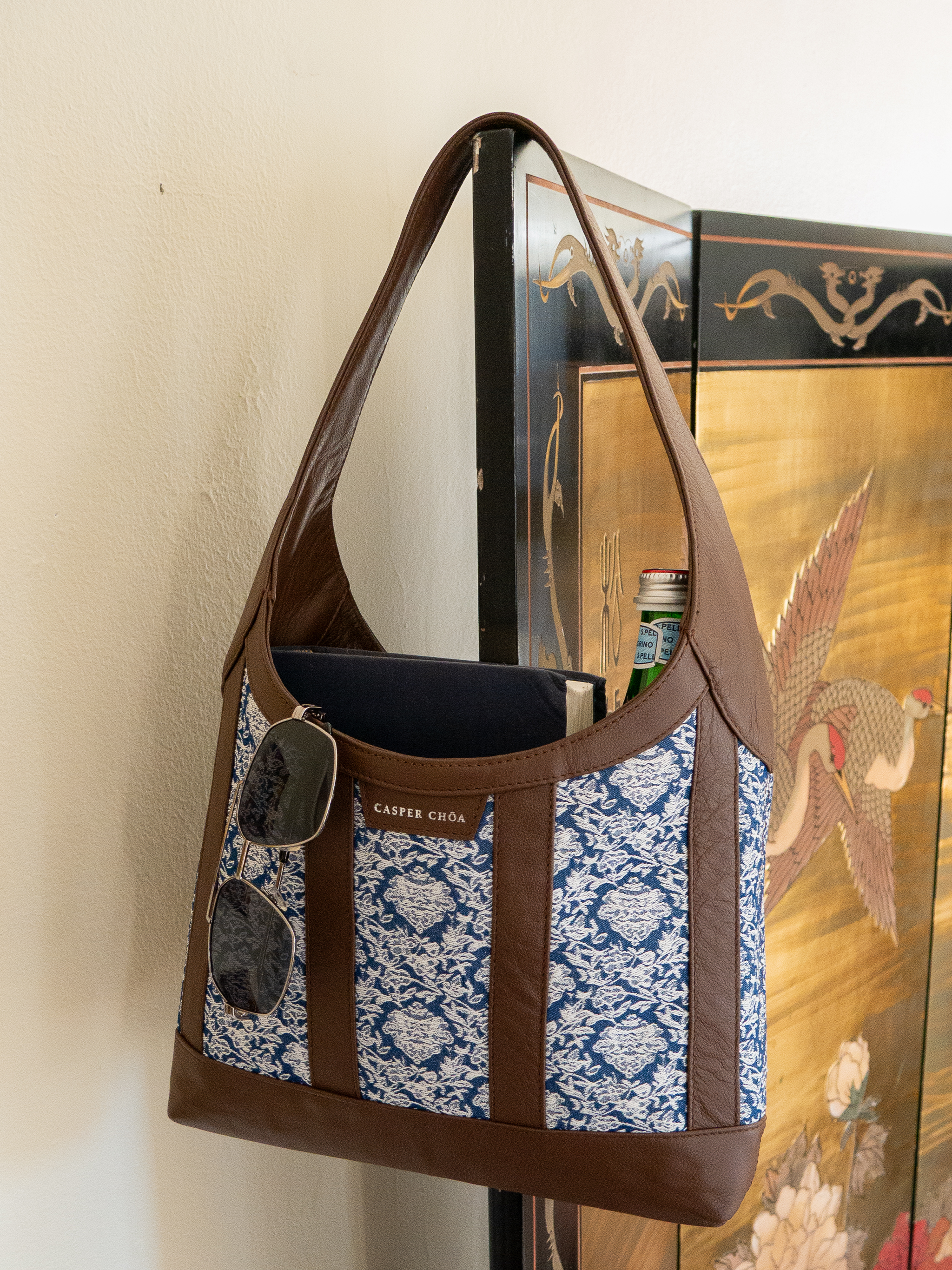 Sunglasses, books, and glass bottle in a brown leather handbag with blue/white floral jacquard against a decorative room divider