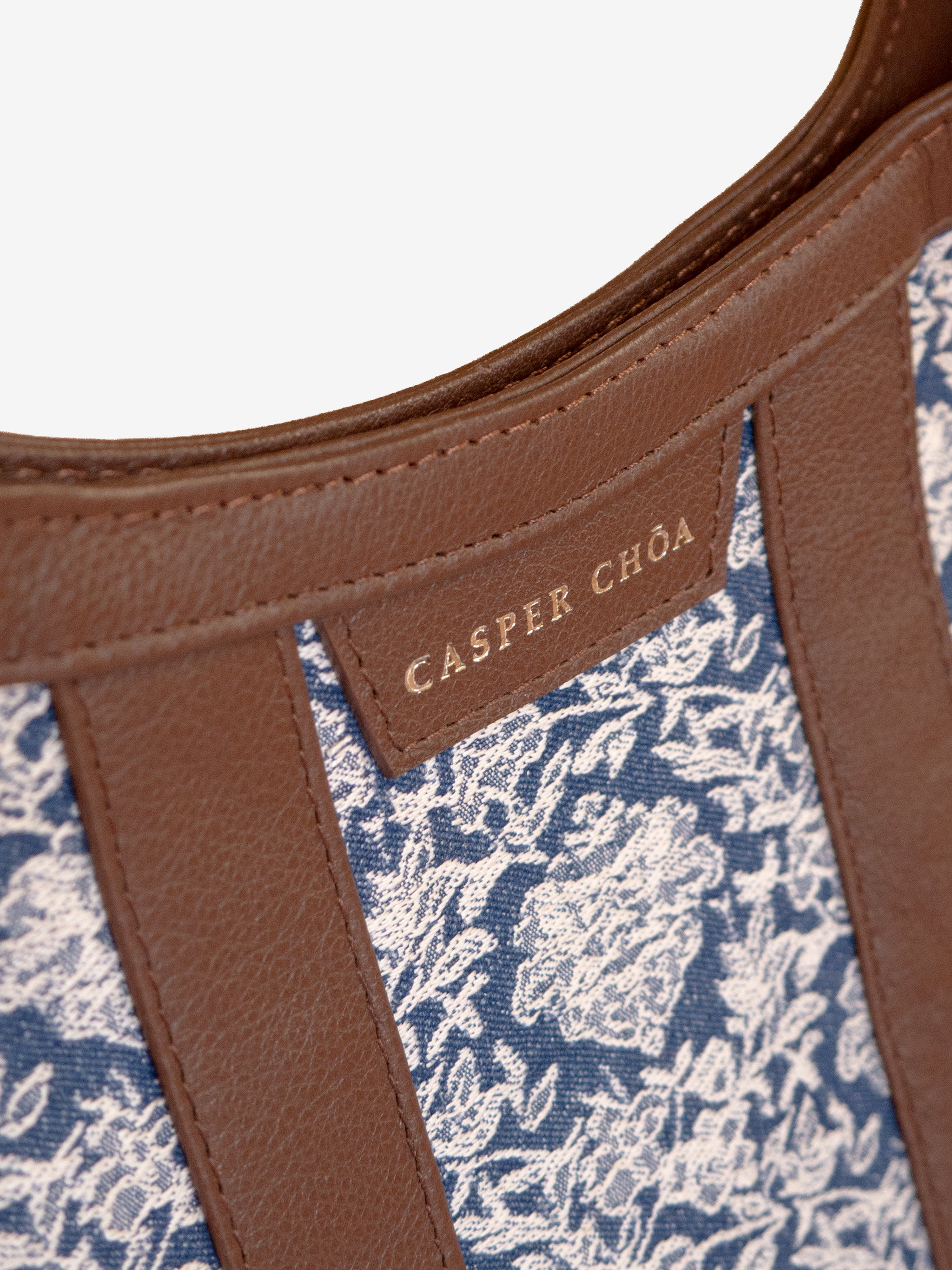 Close-up of a silver-tone CASPER CHOA stamped signature on a brown leather handbag with blue/white floral jacquard on a white background